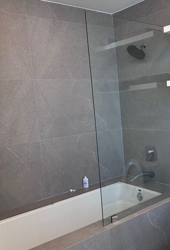 A grey tiled tub and shower with a glass panel