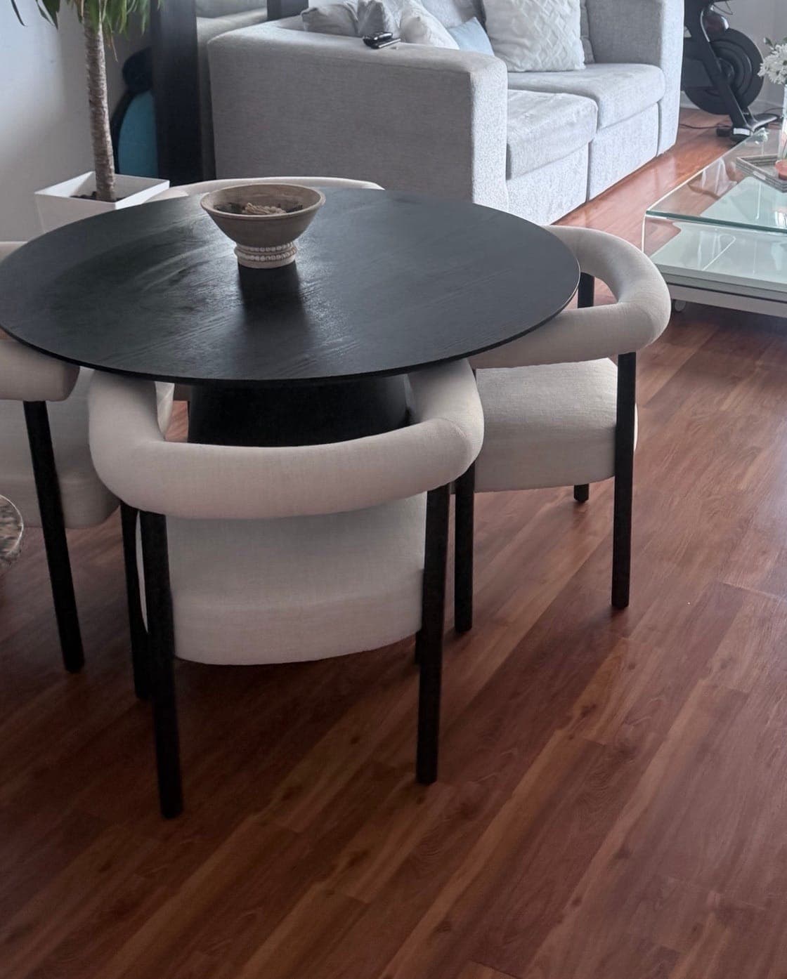 A round black dining table with four cream chairs on a polished wood floor, a grey sofa behind it
